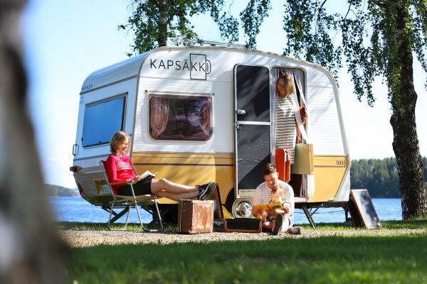 Nelihenkinen perhe hengailee asuntovaunun edessä. Asuntovaunussa lukee Kapsäkki.
