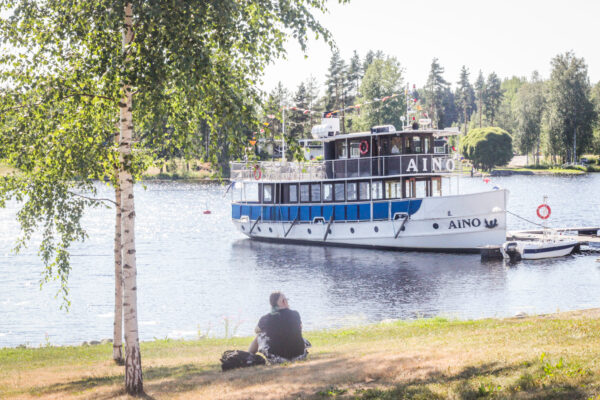 maassa istuva henkilö katselee laiturille päin, johon on kiinnitetty höyrylaiva m/s Aino