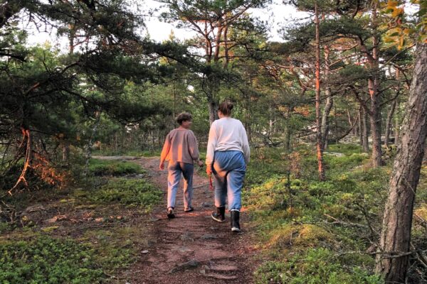Kuvassa kaksi henkilöä kävelevät metsäpolkua pitkin. Kuvassa paistaa ilta-aurinko.